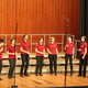 Enthusiastisches Vokalensemble mit 9 Sängerinnen in roten T-Shirts und schwarzen Hosen auf einer Bühne.
